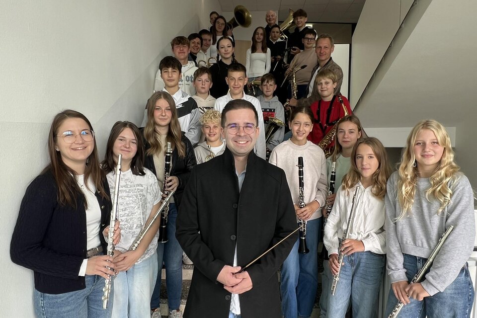 Eine Gruppe von Schülern steht auf einer Treppe und hält Musikinstrumente. Im Vordergrund steht ein Lehrer oder Dirigent, der die Gruppe anlächelt. | © Musikschule Bad Radkersburg