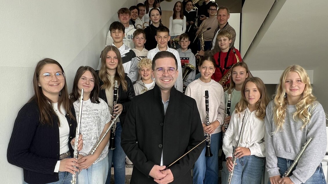 Eine Gruppe von Schülern steht auf einer Treppe und hält Musikinstrumente. Im Vordergrund steht ein Lehrer oder Dirigent, der die Gruppe anlächelt. | © Musikschule Bad Radkersburg