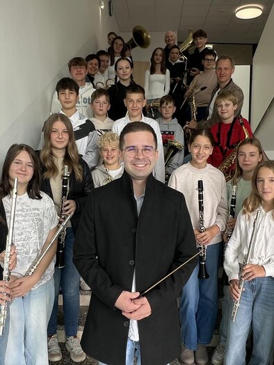 Eine Gruppe von Schülern steht auf einer Treppe und hält Musikinstrumente. Im Vordergrund steht ein Lehrer oder Dirigent, der die Gruppe anlächelt. | © Musikschule Bad Radkersburg