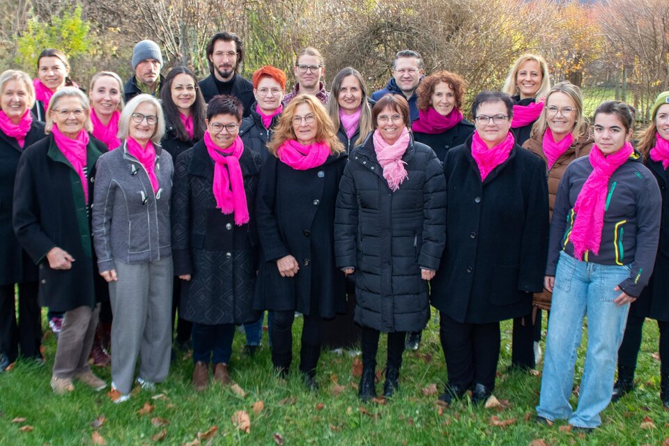 Eine Gruppe von Menschen steht auf einer Wiese, umgeben von Bäumen. Alle tragen schwarze Kleidung und auffällige pinke Schals. | © Singkreis Stainach
