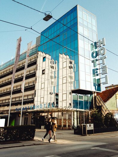 Modern glass facade of the Augarten Art Hotel on a street in Graz with entrance area and hotel signage. | © Mikhail Kyrillos