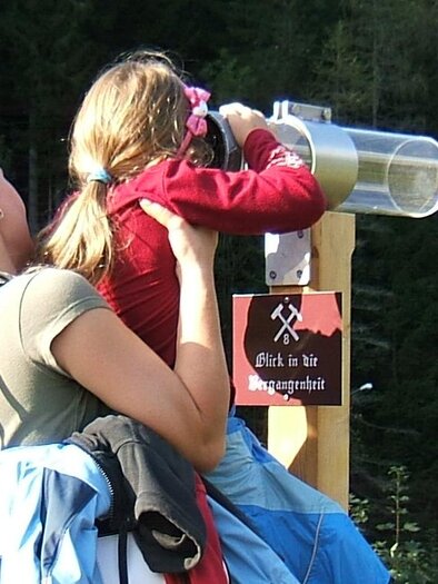Eine Frau hebt ein Kind an, das durch ein Fernrohr schaut. Im Hintergrund sind Bäume und eine ruhige Landschaft zu sehen. | © Naturpark Mürzer Oberland