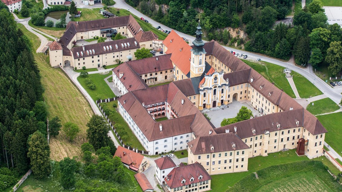 Das Bild zeigt das Stift Rein aus der Vogelperspektive, umgeben von grüner Landschaft am Rande von Graz. Im Zentrum steht die Barockkirche mit ihrem hohen Turm, umgeben von Klostergebäuden und einem Innenhof. Die gelben und weißen Fassaden sowie die roten Dächer kontrastieren mit den umliegenden Wäldern und Wiesen. Das Stift Rein, gegründet 1129, spiegelt den historischen und kulturellen Reichtum der Region wider. | © Region Graz-Harry Schiffer