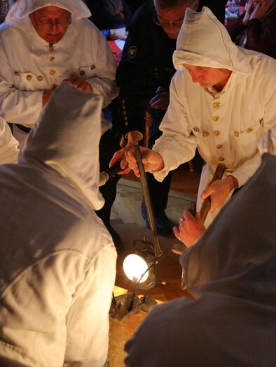 Eine Gruppe von Personen in weißen Kapuzenkleidern versammelt sich um einen Tisch. Sie scheinen an einer Zeremonie oder Ritual beteiligt zu sein, während sie ein Licht betrachten. | © Kulturquartier Leoben