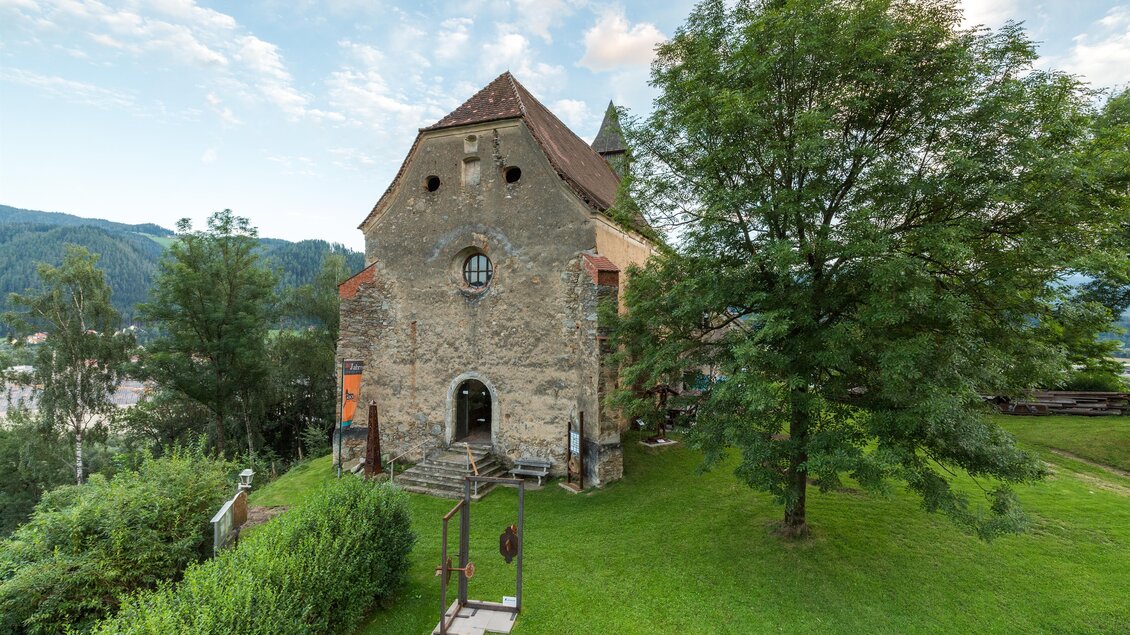 Ein historisches Gebäude steht auf einer grünen Wiese, umgeben von Bäumen. Im Hintergrund sind sanfte Hügel und ein blauer Himmel zu sehen. | © Fotodesign by MarcusAuer