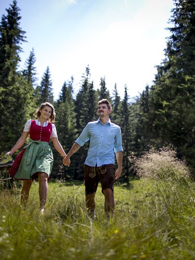 Ein Paar geht Hand in Hand durch eine grüne Wiese im Wald. Die Sonne scheint und der Himmel ist blau. | © Tom Lamm