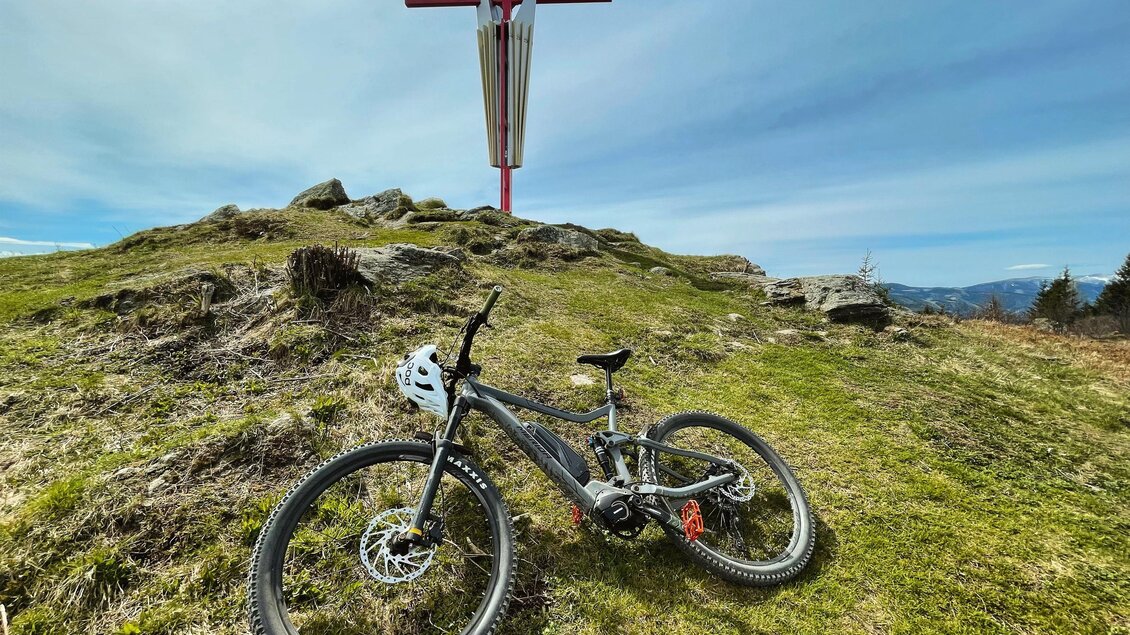 Ein Mountainbike liegt auf einer grünen Wiese. Im Hintergrund steht ein großes Kreuz vor einem klaren Himmel. | © Manuel Stadler