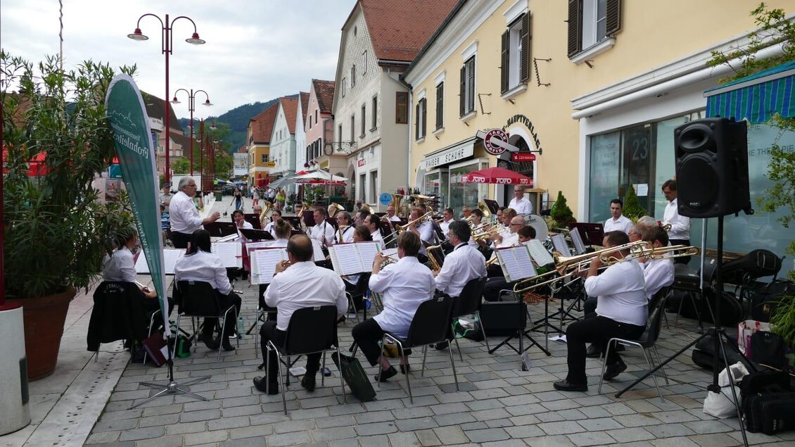 Eine Blaskapelle spielt im Freien auf einer belebten Straße. Im Hintergrund sind zahlreiche Häuser und Sitzgelegenheiten zu sehen. | © WOKA