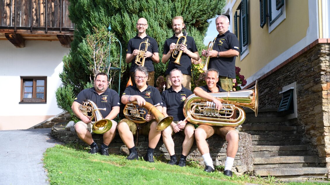 Eine Gruppe von Musikern steht im Freien und spielt Blechblasinstrumente. Im Hintergrund sind Bäume und ein Gebäude zu sehen. | © (c) KK