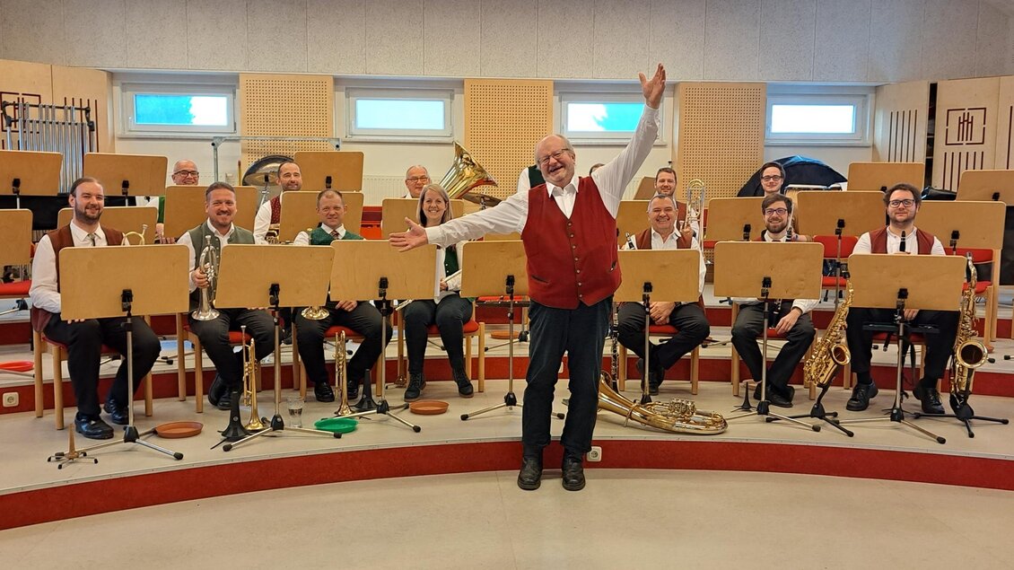 Eine Gruppe von Musikern steht auf der Bühne und spielt verschiedene Blasinstrumente. Im Vordergrund lächelt ein Dirigent begeistert.