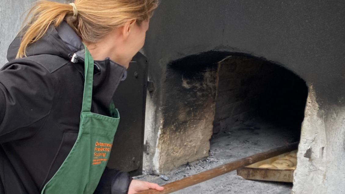 <p>Eine Frau schiebt Brotteig mit Holzschieber in historischen Brotbackofen im Österreichischen Freilichtmuseum Stübing.</p> | © ÖFM