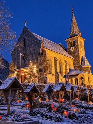 Eine historische Kirche im Schnee bei Dämmerung. Der Garten ist mit Laternen und Schneeverzierungen geschmückt. | © Bettina Scheck