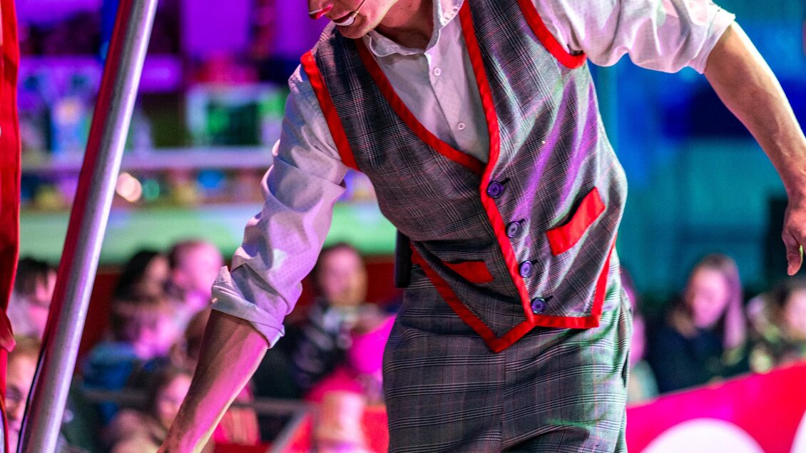 Ein Zirkusmitarbeiter in einem farbenfrohen Kostüm steht in der Manege. Er hält ein Spielzeug-Löwe in der Hand und hat ein Lächeln auf dem Gesicht. | © Circus Aros