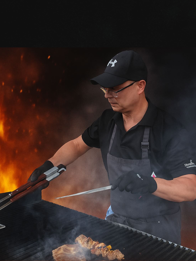 Ein Mann grillt mit Freude Fleisch auf einem großen Grill. Im Hintergrund sind Flammen und Rauch sichtbar. | © Hotel Domittner