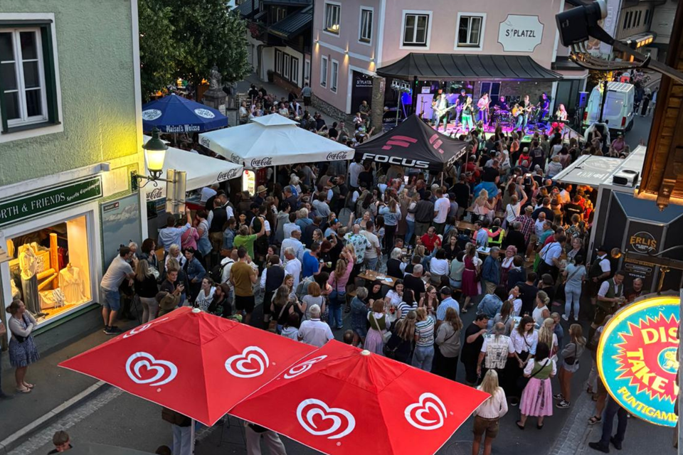 Eine große Menschenmenge versammelt sich auf einer Straße, um eine Live-Veranstaltung zu genießen. Rote Sonnenschirme und bunte Lichter schaffen eine festliche Atmosphäre. | © Roland Kasperer