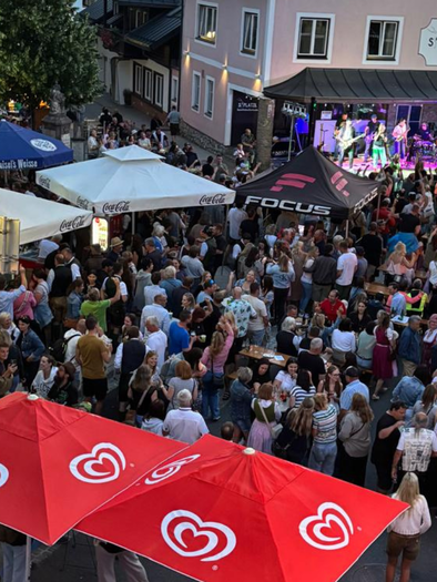 A large crowd gathers on a street to enjoy a live event. Red sun umbrellas and colorful lights create a festive atmosphere. | © Roland Kasperer