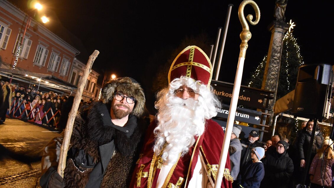 Zwei Männer in festlichen Kostümen stehen bei Nacht zusammen. Der eine trägt einen langen weißen Bart und eine bischöfliche Mitra, während der andere einen pelzbesetzten Umhang und einen Stock hat. | © Foto de Monte Mureck