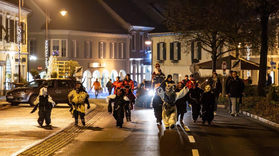 Eine Gruppe von Menschen in festlichen Kostümen zieht nachts eine Straße entlang. Die Umgebung ist beleuchtet und es sind einige Autos zu sehen. | © Foto de Monte Mureck