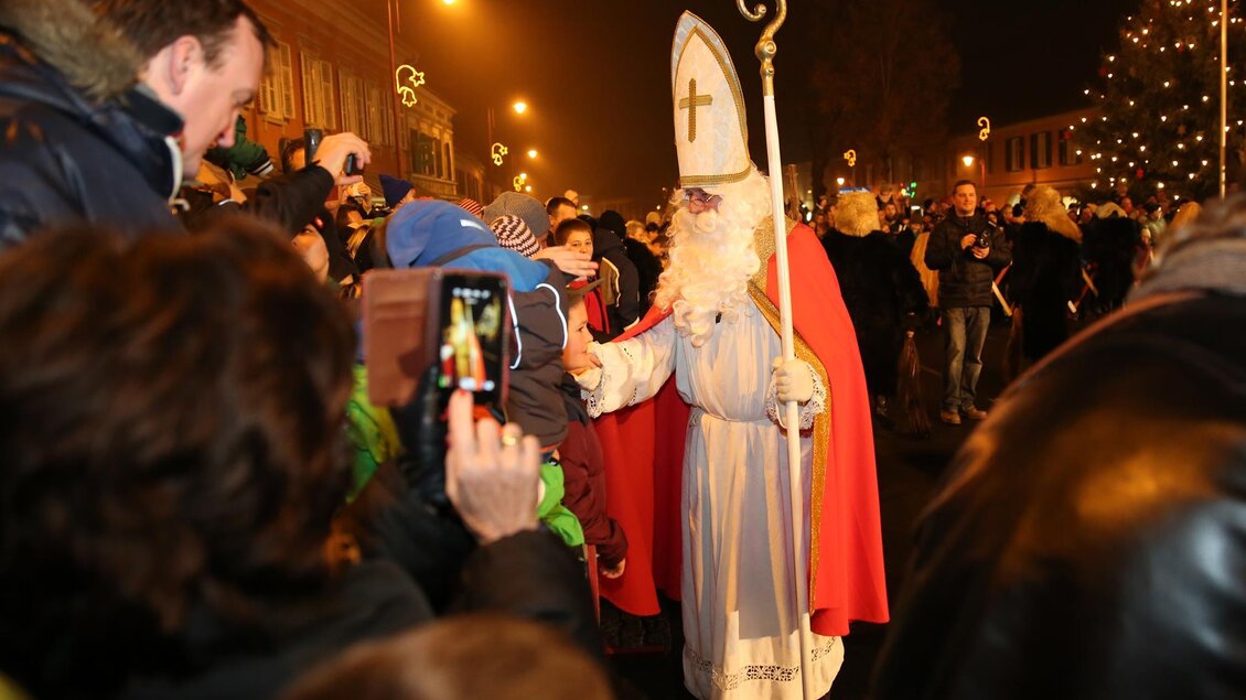 Eine festliche Menschenmenge empfängt den heiligen Nikolaus, der in einem langen, weißen Gewand und mit einem roten Umhang auftritt. Der Nikolaus interagiert freundlich mit den Kindern und hält einen Stab in der Hand. | © Foto de Monte Mureck