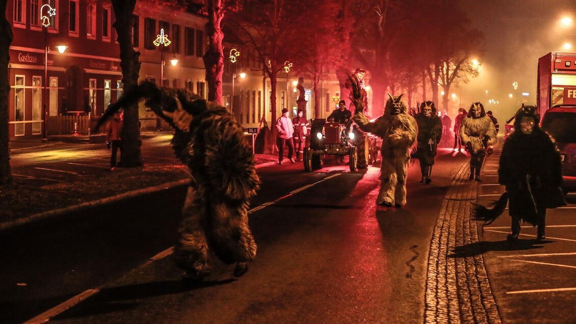 Eine schaurige Nachtparade mit maskierten Teilnehmern in unheimlichen Kostümen. Die Straße ist schwach beleuchtet und mit rotem Licht durchzogen. | © Foto de Monte Mureck