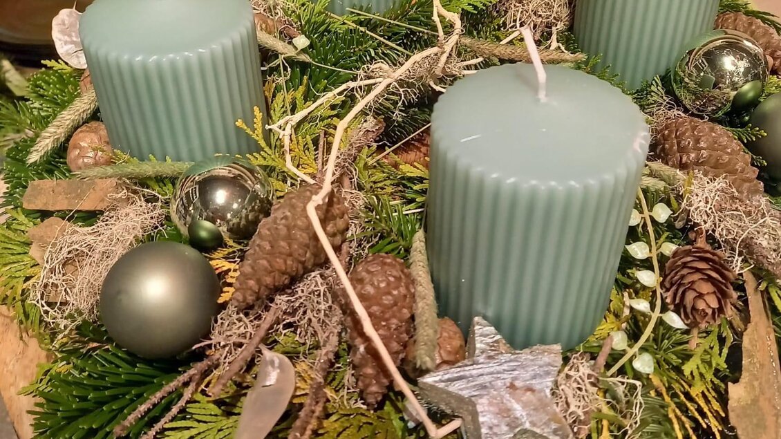 Ein schön dekorierter Adventskranz mit vier blauen Kerzen, umgeben von Tannenzweigen, Zapfen und glitzernden Ornamenten. Die Anordnung strahlt eine gemütliche und festliche Atmosphäre aus. | © Blumenhof Bender