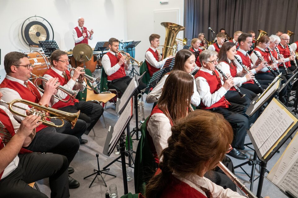 Eine Blaskapelle spielt in einem Saal. Die Musiker tragen rote Westen und spielen verschiedene Blasinstrumente. | © echtzeit-tv, Stefan Lendl.