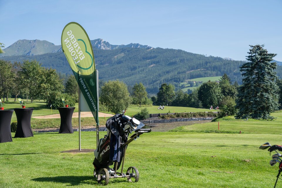 Eine malerische Golfplatzlandschaft mit Bergen im Hintergrund. Im Vordergrund steht ein Golfwagen und ein grünes Veranstaltungsbanner. | © K.Paller