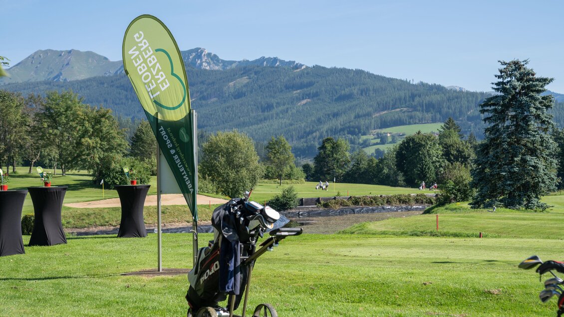 Eine malerische Golfplatzlandschaft mit Bergen im Hintergrund. Im Vordergrund steht ein Golfwagen und ein grünes Veranstaltungsbanner. | © K.Paller