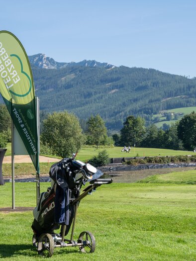 Eine malerische Golfplatzlandschaft mit Bergen im Hintergrund. Im Vordergrund steht ein Golfwagen und ein grünes Veranstaltungsbanner. | © K.Paller
