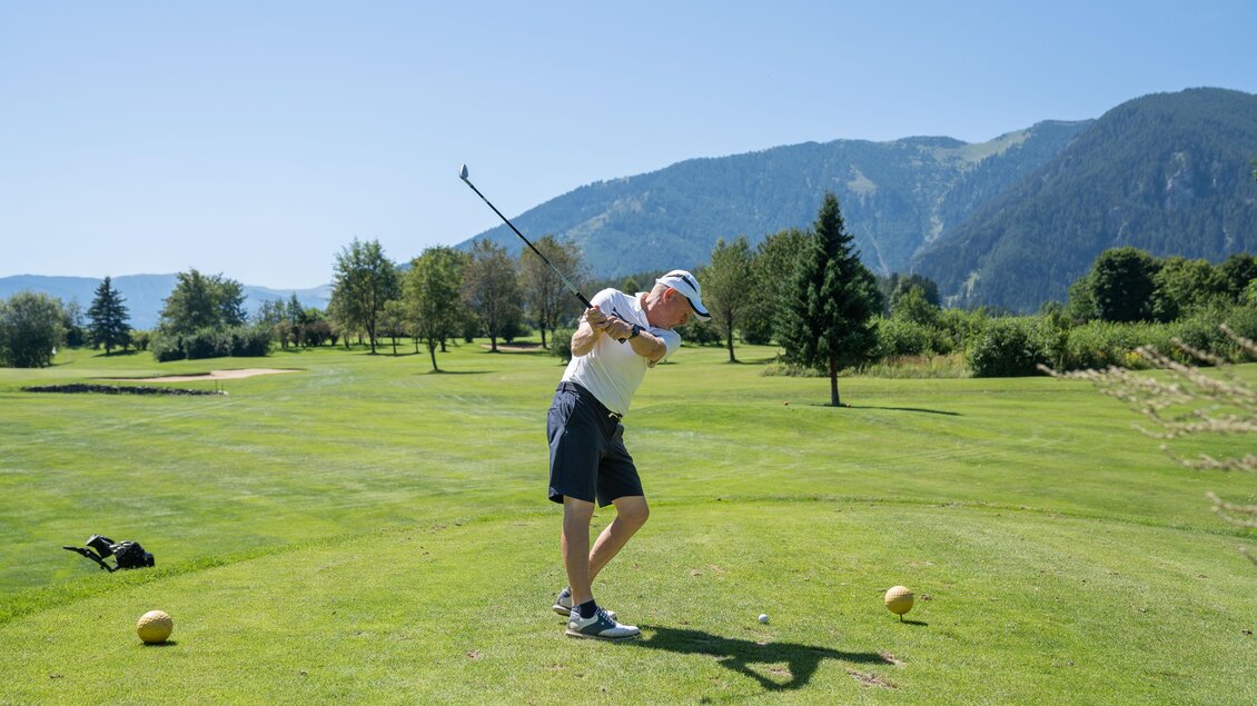 Ein Golfer steht auf einem Abschlagplatz und macht einen Schwung. Im Hintergrund sind Bäume und Berge zu sehen. | © K.Paller