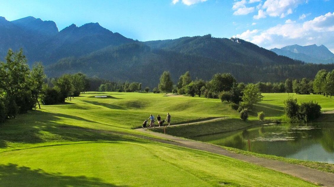 Eine malerische Golfanlage mit grünen Fairways und sanften Hügeln. Im Hintergrund sind majestätische Berge und ein ruhiger Teich zu sehen. | © Foto Freisinger