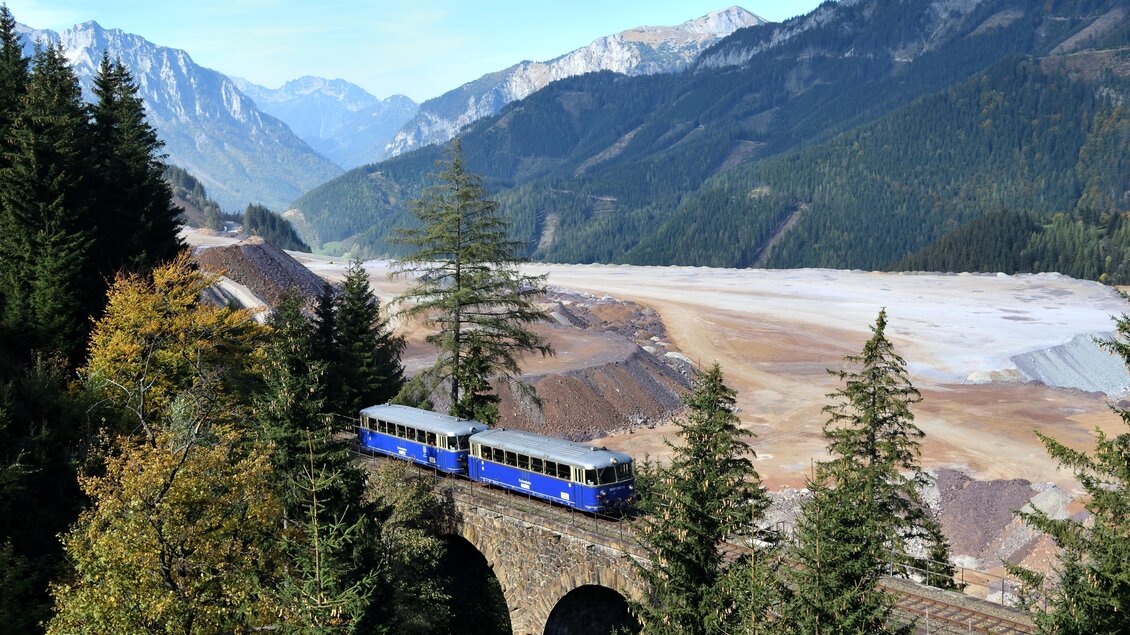 Ein blaublühender Zug fährt über eine historische Brücke inmitten einer malerischen Berglandschaft. Im Hintergrund sind hohe Berge und ein weites, schottriges Tal zu sehen. | © Erzbergbahn