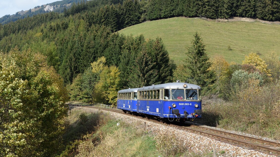 Ein blauer Schienenbus fährt durch eine malerische Landschaft mit Bäumen und Hügeln. Die Sonne scheint und das Wetter ist klar. | © Verein Erzbergbahn