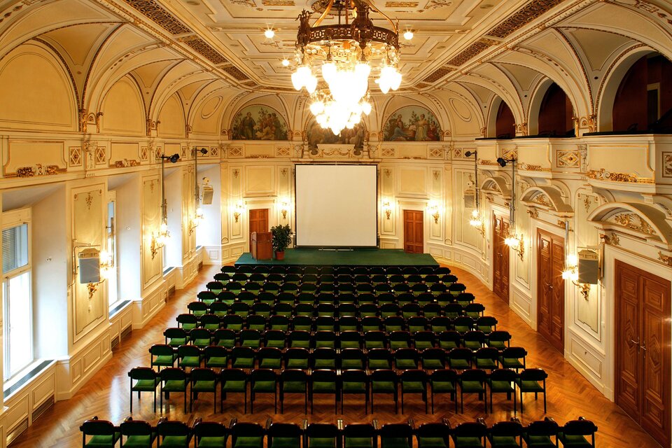 Ein eleganter Veranstaltungsraum mit vielen grünen Stühlen und einer großen Leinwand. Die Decke ist kunstvoll gestaltet und es hängt ein großer Kronleuchter. | © (c) CG