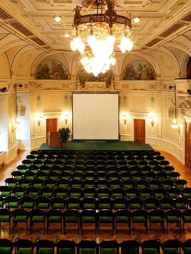 An elegant event space with many green chairs and a large screen. The ceiling is intricately designed, and a large chandelier hangs from it. | © (c) CG