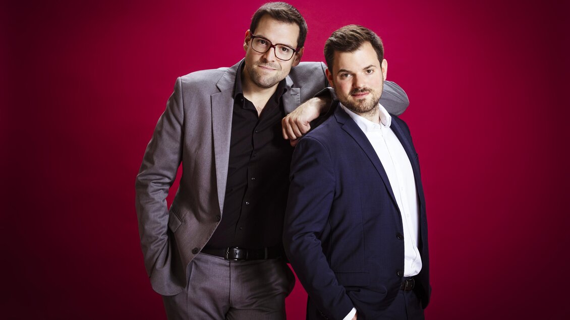 Zwei Herren in eleganter Kleidung stehen vor einem pinken Hintergrund. Sie lächeln und wirken freundlich und entspannt. | © Moritz Schell
