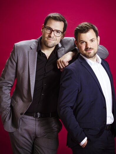 Zwei Herren in eleganter Kleidung stehen vor einem pinken Hintergrund. Sie lächeln und wirken freundlich und entspannt. | © Moritz Schell