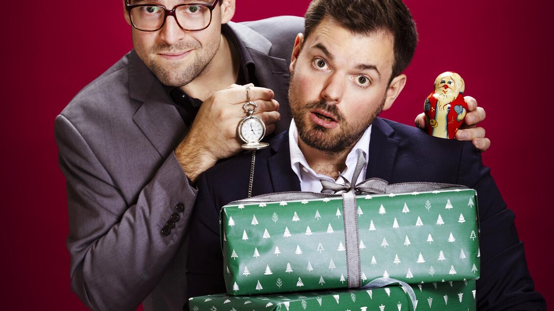 Zwei Männer stehen vor einem roten Hintergrund und halten Geschenke in grüner Geschenkverpackung. Einer der Männer trägt eine Brille und hat eine lebhafte Mimik. | © Moritz Schell