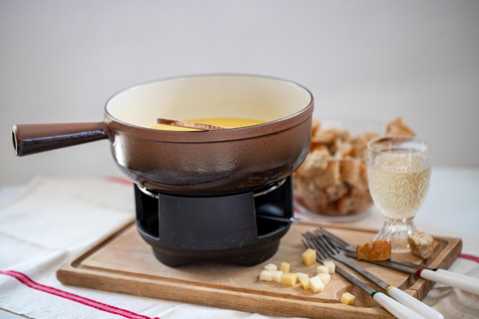 Eine Käsefondue-Pfanne steht auf einem Tisch, umgeben von Brotstücken und einem Glas Wein. Das Fondue hat eine goldene Farbe und wird mit einem Holzlöffel serviert. | © Pixabay