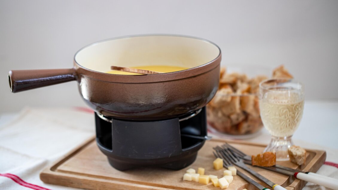 Eine Käsefondue-Pfanne steht auf einem Tisch, umgeben von Brotstücken und einem Glas Wein. Das Fondue hat eine goldene Farbe und wird mit einem Holzlöffel serviert. | © Pixabay