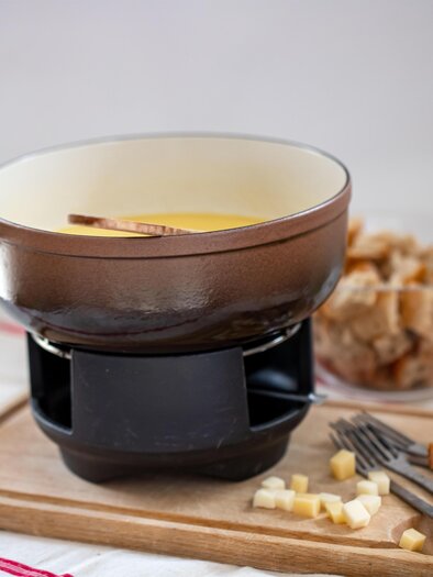 Eine Käsefondue-Pfanne steht auf einem Tisch, umgeben von Brotstücken und einem Glas Wein. Das Fondue hat eine goldene Farbe und wird mit einem Holzlöffel serviert. | © Pixabay