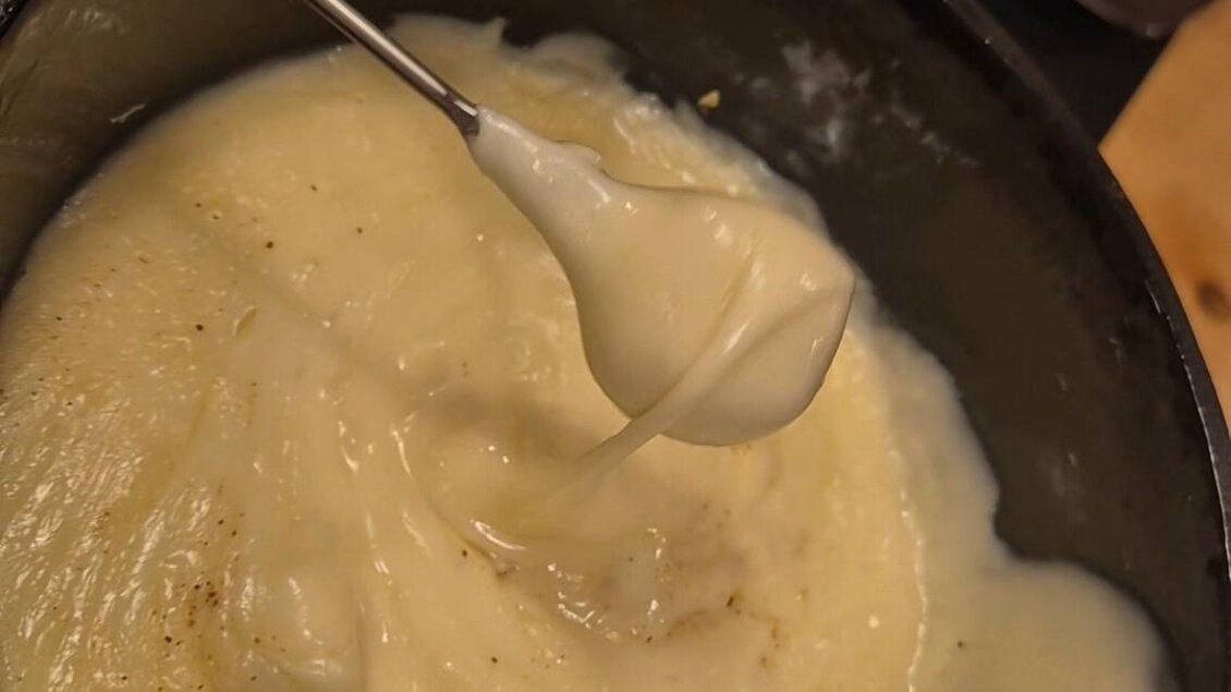 Ein Topf mit cremigem Käsefondue und einem Löffel. Im Hintergrund ist ein Korb mit Brot zu sehen. | © Almenland Stollenkäse
