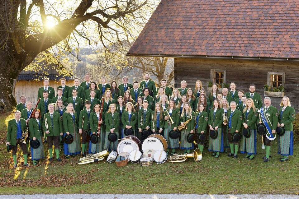 Eine große Musikgruppe in traditioneller bayerischer Tracht steht auf einer Bühne. Die Mitglieder halten verschiedene Musikinstrumente und lächeln in die Kamera. | © MMK Passail_Oststeiermark
