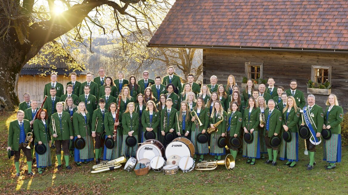 Eine große Musikgruppe in traditioneller bayerischer Tracht steht auf einer Bühne. Die Mitglieder halten verschiedene Musikinstrumente und lächeln in die Kamera. | © MMK Passail_Oststeiermark