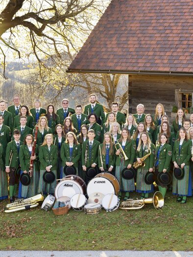 Eine große Musikgruppe in traditioneller bayerischer Tracht steht auf einer Bühne. Die Mitglieder halten verschiedene Musikinstrumente und lächeln in die Kamera. | © MMK Passail_Oststeiermark