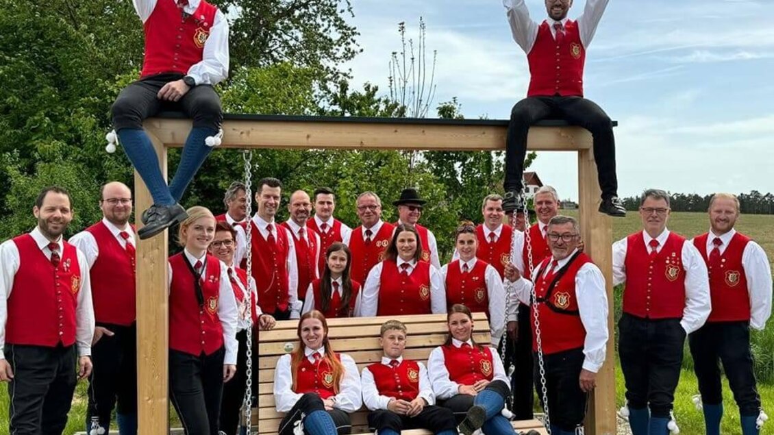 Eine große Blasmusikgruppe posiert auf einer Wiese vor einer Steinmauer. Alle Mitglieder tragen traditionelle Trachten und halten verschiedene Musikinstrumente. | © Trachtenkapelle Ebersdorf_Oststeiermark