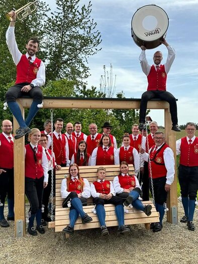 Eine große Blasmusikgruppe posiert auf einer Wiese vor einer Steinmauer. Alle Mitglieder tragen traditionelle Trachten und halten verschiedene Musikinstrumente. | © Trachtenkapelle Ebersdorf_Oststeiermark