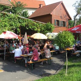 A cheerful outdoor restaurant with many guests seated at tables. Colorful umbrellas provide shade under the green arbor. | © Ga Weint Gehn Gemeinschaft