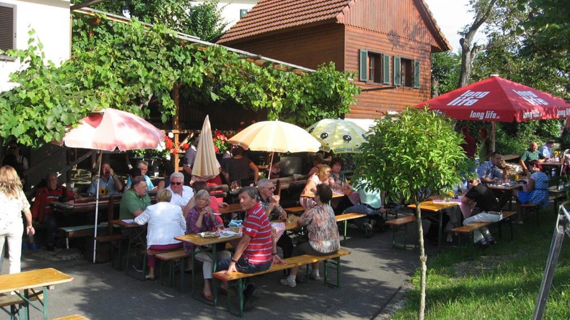 Ein fröhliches Restaurant im Freien mit vielen Gästen, die an Tischen sitzen. Bunte Schirme spenden Schatten unter der grünen Laube. | © Ga Weint Gehn Gemeinschaft