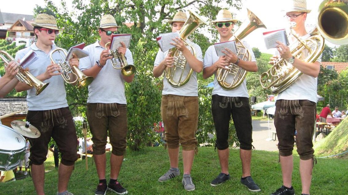 Fünf Musiker spielen mit verschiedenen Blechblasinstrumenten im Freien. Sie tragen Hütchen und Sonnenbrillen und stehen auf einer grünen Wiese. | © Ga Weint Gehn Gemeinschaft